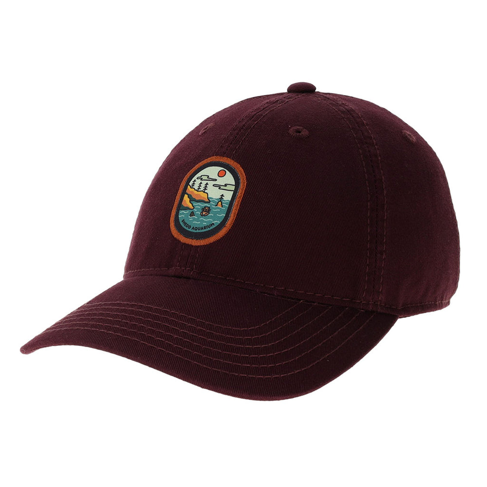The Shedd Aquarium Cap is a maroon baseball hat with a colorful embroidered patch of mountains, trees, water, and sun on the front, inspired by vibrant aquatic life.
