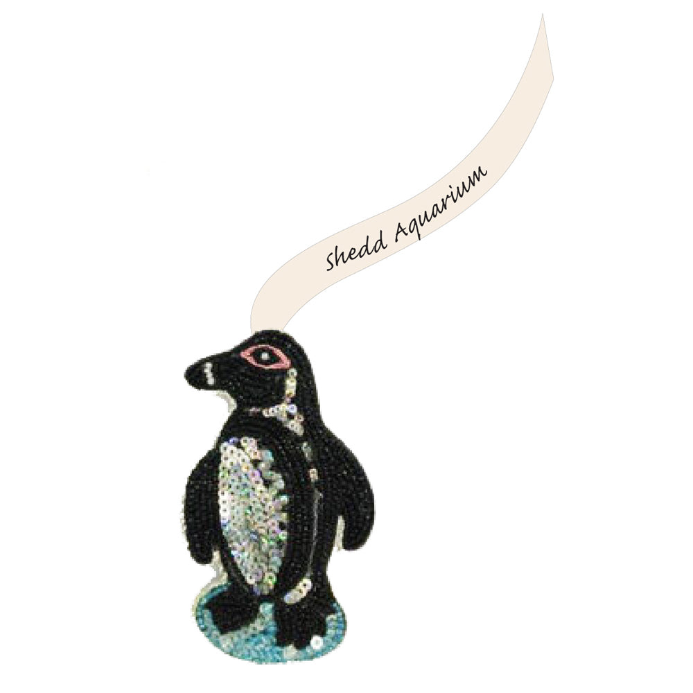 The Sequin Penguin Ornament stands on a blue base with a cream ribbon reading "Shedd Aquarium"—an ideal, unique addition to your holiday decor.