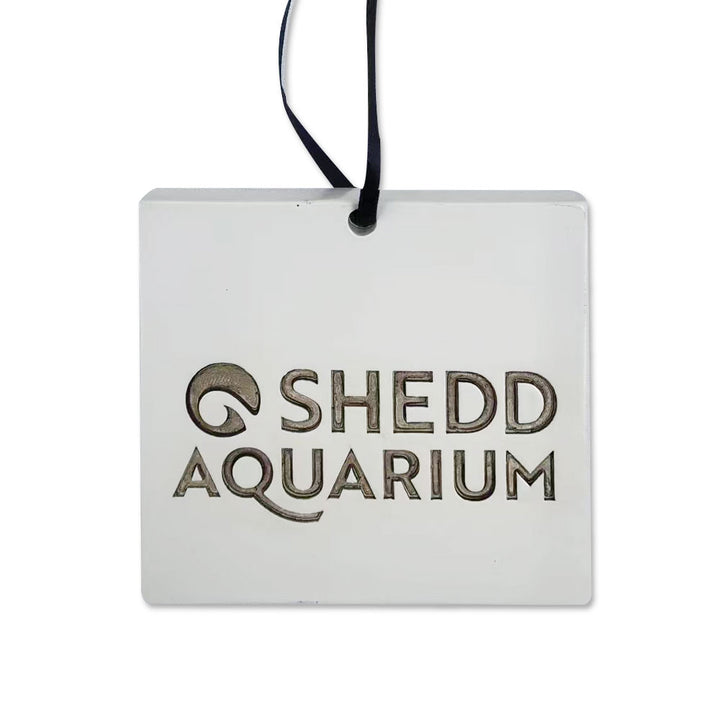 The Shedd Aquarium Ceiling Tile Ornament is a white rectangular ornament with a wave design, metallic-engraved lettering, and a black ribbon—perfect as an aquarium gift or unique ceiling tile accent.