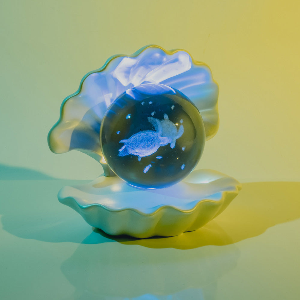 The Sea Life Etched Glass Globe, showcasing a sea turtle, sits in an open ceramic clam shell with gentle yellow and blue lighting—an elegant marine life accent.