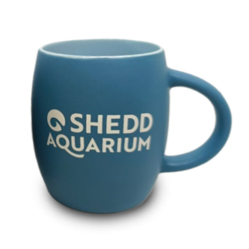 The Shedd Aquarium Penguin Etched Mug is a blue ceramic mug with a large handle, featuring an etched penguin design alongside the white Shedd Aquarium logo and text on its side. Set against a plain white background, this mug is an ideal keepsake for marine life enthusiasts.