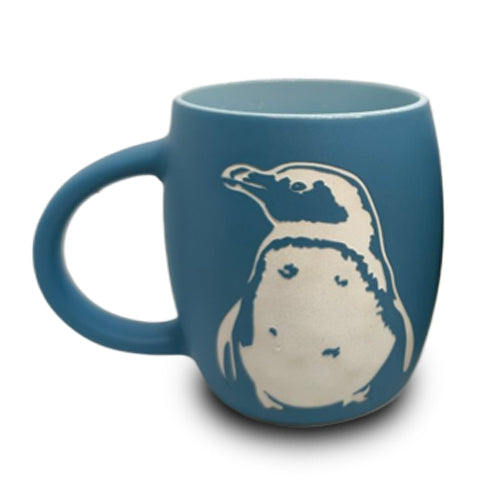 The Shedd Aquarium Penguin Etched Mug is a blue ceramic mug with a large handle, featuring a white outline drawing of a penguin on its side—celebrating marine life and inspired by the wonders of the Shedd Aquarium.