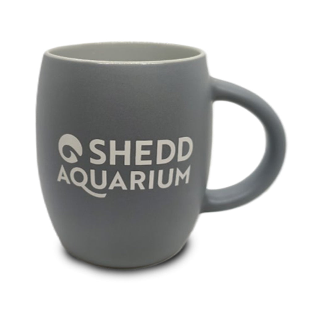 Introducing the Shedd Aquarium Beluga Etched Mug—a gray ceramic mug with a handle, featuring “Shedd Aquarium” in white lettering on the front. This unique mug is ideal for marine life conservation enthusiasts or anyone looking for a distinctive beluga whale-themed drinkware item.