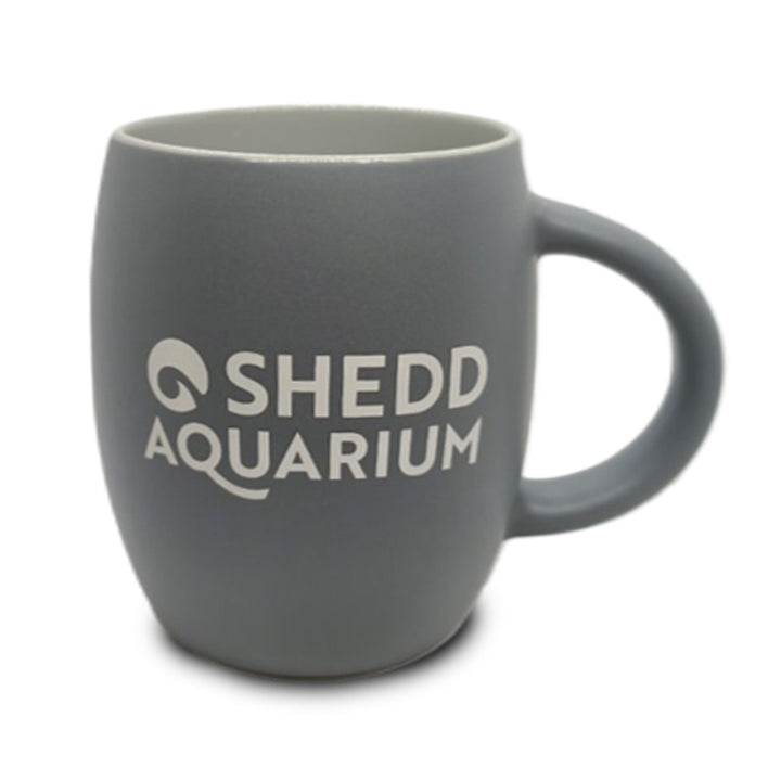 Introducing the Shedd Aquarium Beluga Etched Mug—a gray ceramic mug with a handle, featuring “Shedd Aquarium” in white lettering on the front. This unique mug is ideal for marine life conservation enthusiasts or anyone looking for a distinctive beluga whale-themed drinkware item.