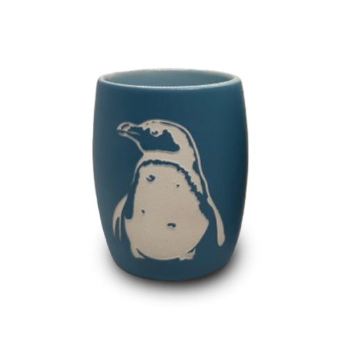 The Shedd Aquarium Penguin Etched Shot Glass is a blue ceramic cup featuring a white outline illustration of a penguin on the front. With its smooth, rounded shape, this charming wildlife keepsake stands out against a plain white background.