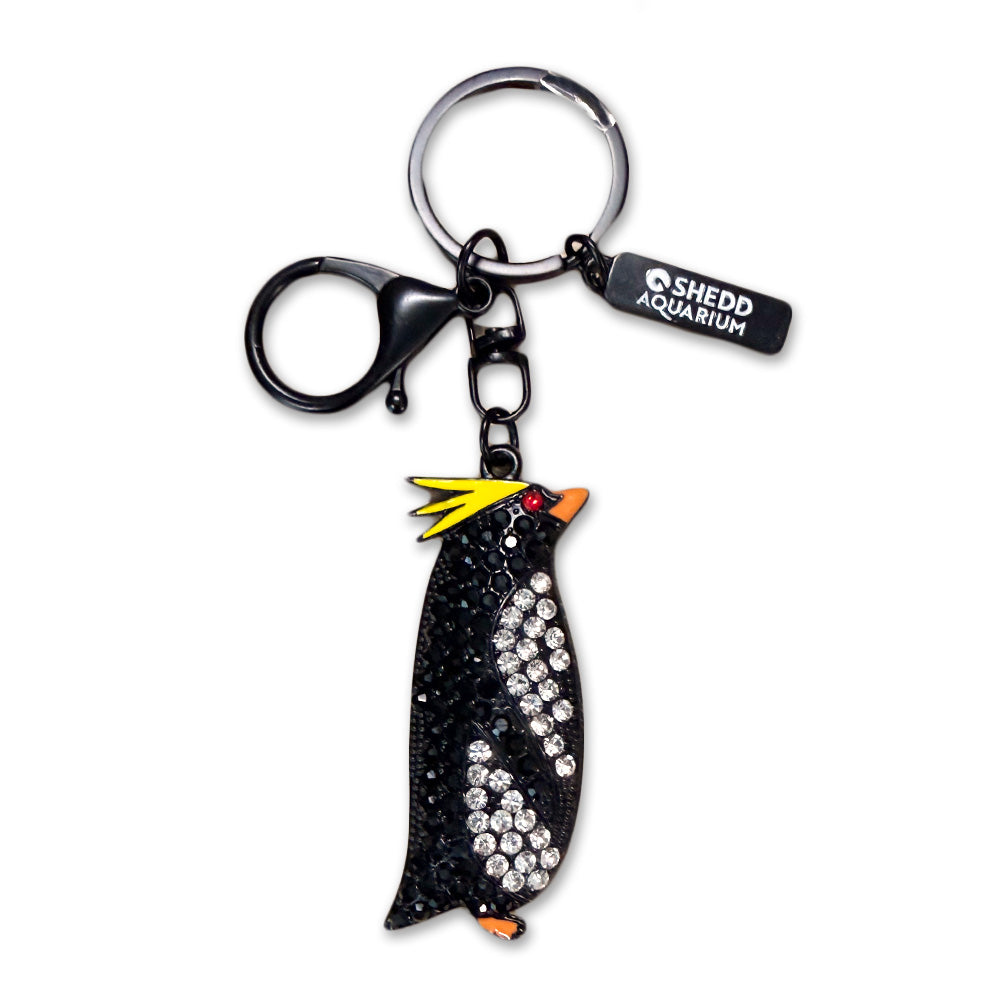 The Shedd Aquarium Rock Hopper Penguin Bling Keychain features sparkling details, yellow head feathers, an orange beak, a black and white body, and comes with a tag labeled Shedd Aquarium.