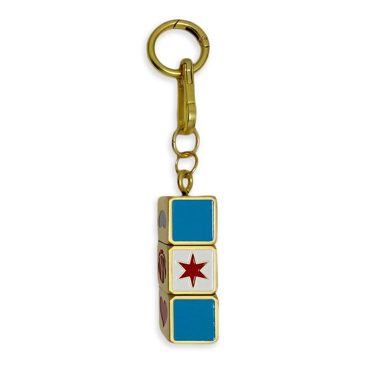 Introducing the Chicago Block Spinner Bag Charm—a gold keychain featuring a small rectangular block adorned with blue squares, a red star, and multicolored icons on its sides. This distinctive accessory brings a playful and vibrant touch to any collection.
