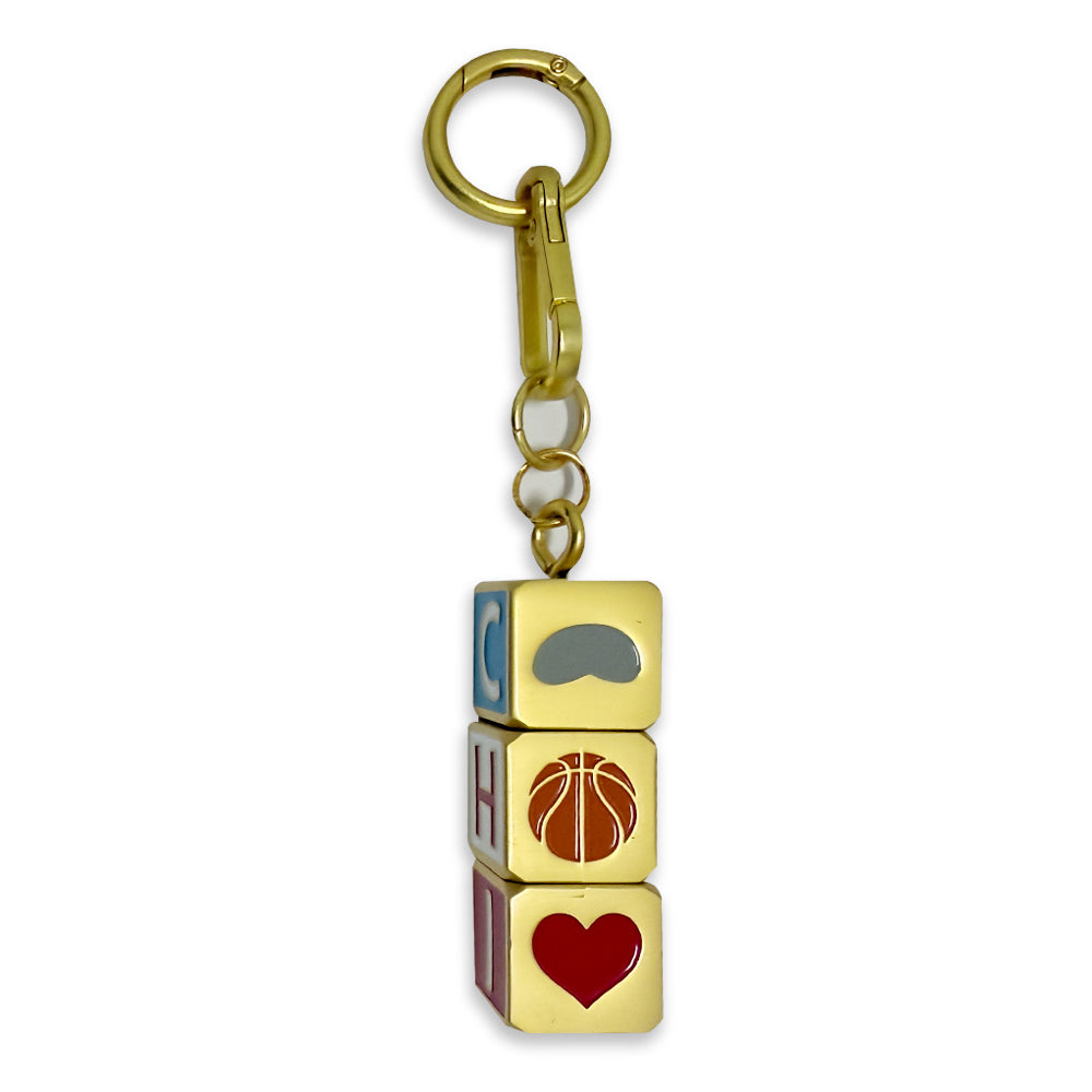 Introducing the Chicago Block Spinner Bag Charm: a gold keychain featuring three stacked square blocks, ideal as a stylish Chicago accessory for your bag. The top block displays a gray mustache, the middle showcases a brown basketball, and the bottom features a red heart. Letters are visible along the sides of each block, making this fun and eye-catching charm the perfect addition to any bag or set of keys.