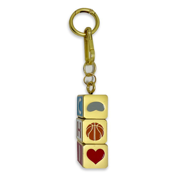 Introducing the Chicago Block Spinner Bag Charm: a gold keychain featuring three stacked square blocks, ideal as a stylish Chicago accessory for your bag. The top block displays a gray mustache, the middle showcases a brown basketball, and the bottom features a red heart. Letters are visible along the sides of each block, making this fun and eye-catching charm the perfect addition to any bag or set of keys.