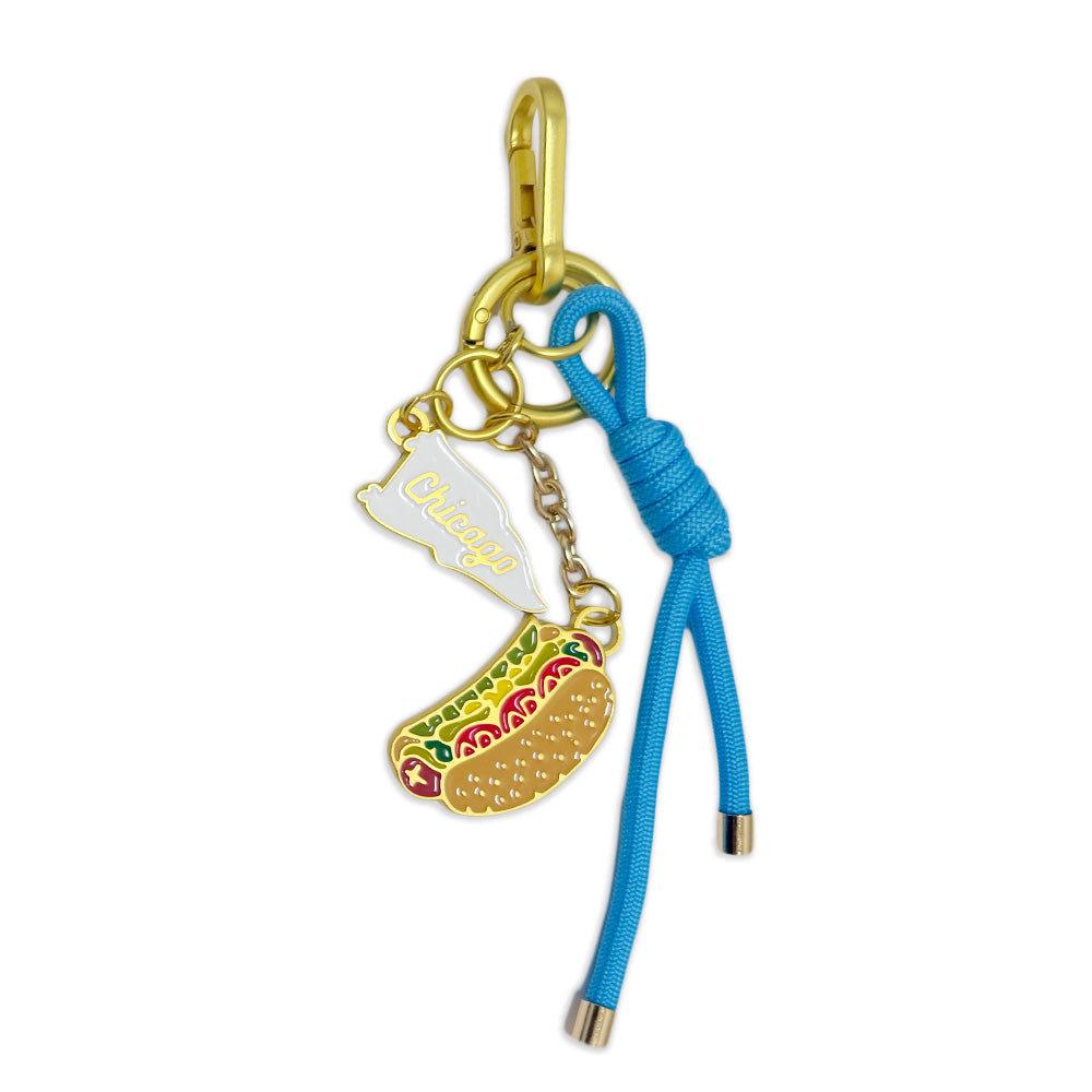 Introducing the Chicago Icons Dangle Bag Charm: this playful accessory features a gold clip, a knotted blue cord, a metal charm shaped like a classic Chicago hot dog, and a white charm with “Chicago” written in yellow cursive. Celebrate the Windy City’s unique spirit and architectural heritage every day with this standout piece inspired by one of America’s most iconic cultural destinations.