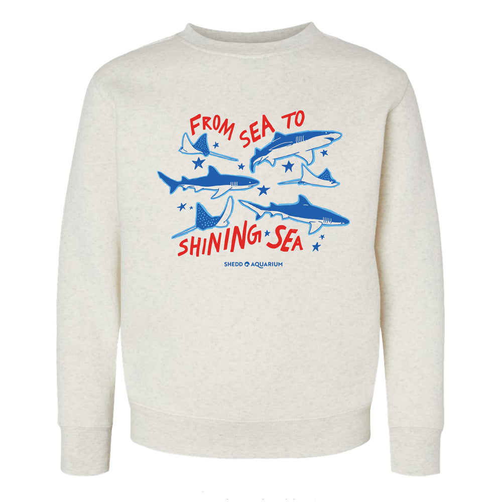 The Shedd Aquarium Sea to Shining Sea Youth Crew Sweatshirt features a cream color, blue shark and ray illustrations, and red and blue "From Sea to Shining Sea" text, finished with the Shedd Aquarium logo at the bottom.