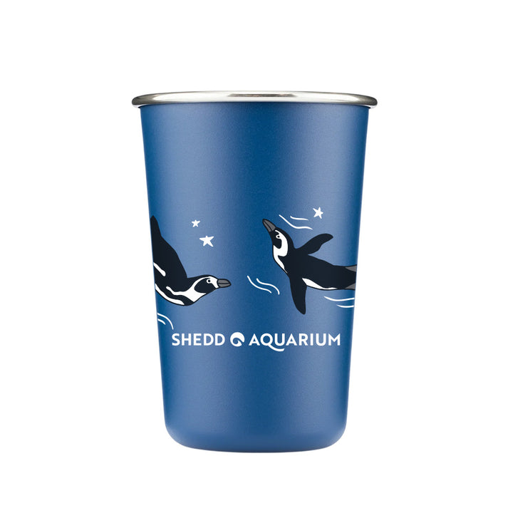 Shedd Aquarium Red White & Ocean Blue Stars Pint Cup: a blue metal cup with swimming penguins, white stars, and waves. “Shedd Aquarium” is printed in white near the bottom—a charming, eco-friendly marine life tribute.