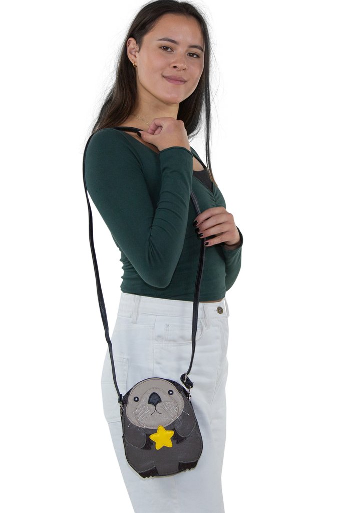 A smiling woman with long dark hair in a dark green long-sleeve top and white pants holds the Crossbody Sea Otter Bag—a cute gray otter bag with a yellow star and shoulder strap—against a white background.