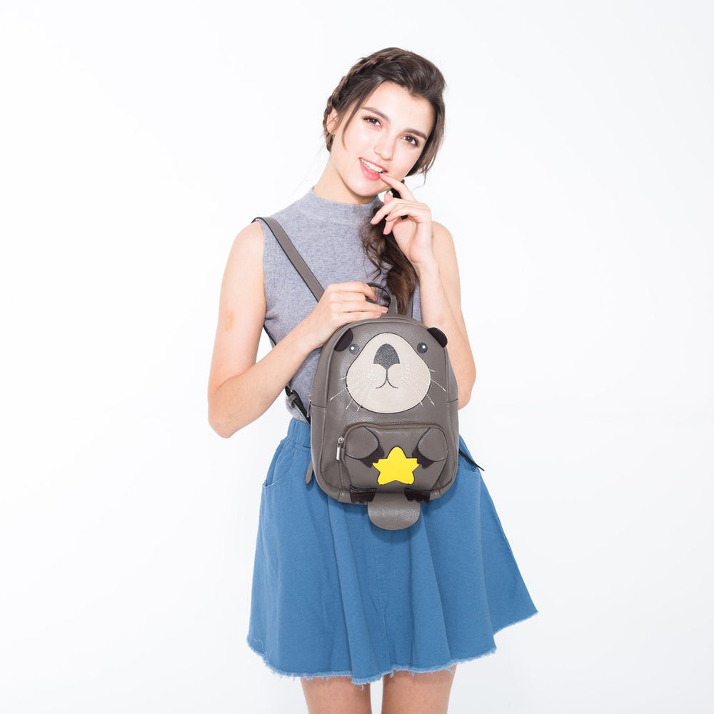 A young woman with braided hair stands against a white background, wearing a gray sleeveless top, blue skirt, and carrying the Mini Sea Otter Backpack featuring a cute bear face and yellow star on the front.