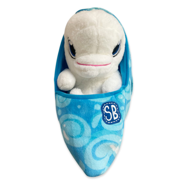 A Swaddle Babies Beluga Plush with blue eyes and a pink mouth sits inside a blue slipper featuring swirl patterns and an "SB" patch, set against a white background.