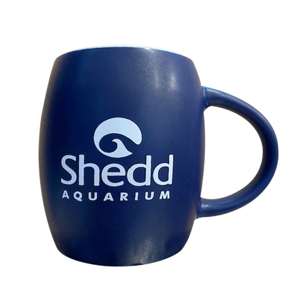 The Blue Shedd Aquarium Penguin Mug is a ceramic mug with a rounded shape and large handle, featuring the Shedd Aquarium logo and name in white on the front.