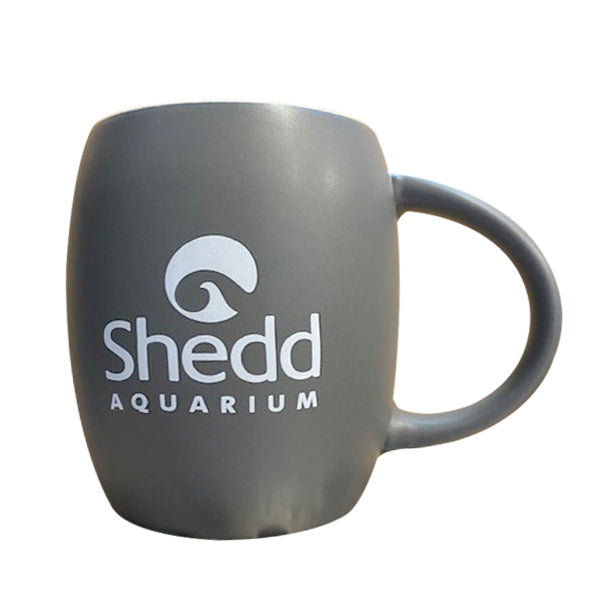 The Grey Shedd Aquarium Beluga Mug is a matte gray ceramic mug featuring the Shedd Aquarium logo and text in white, a rounded shape, and a large handle—perfect for aquarium fans.