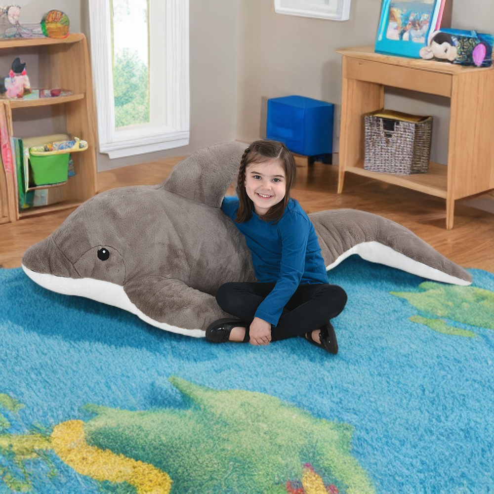 Child sitting on a large shark-shaped pillow in a room with toys and furniture.