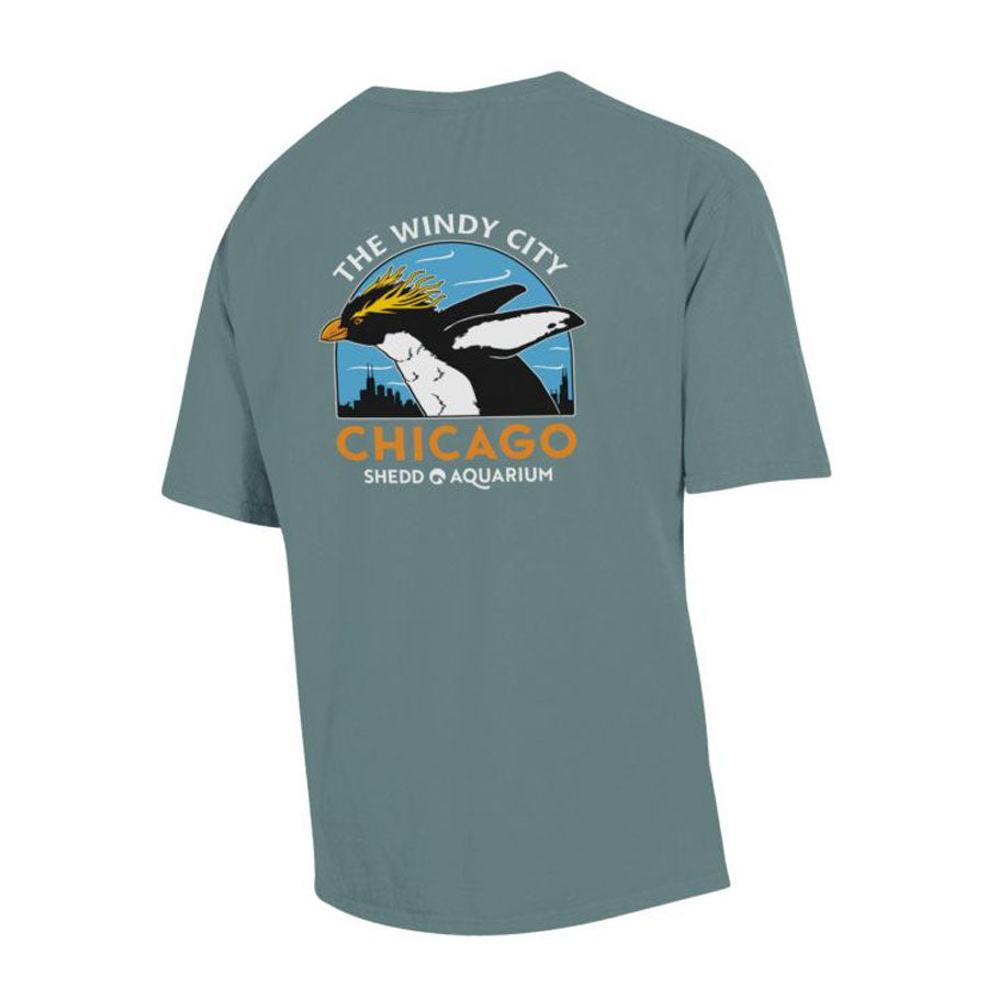 The Windy City T-Shirt is blue, with an orca in a yellow hat and Chicago’s skyline graphic. “THE WINDY CITY” appears above, “CHICAGO SHED AQUARIUM” below. A Brainfreeze Press favorite—perfect for penguin fans!.